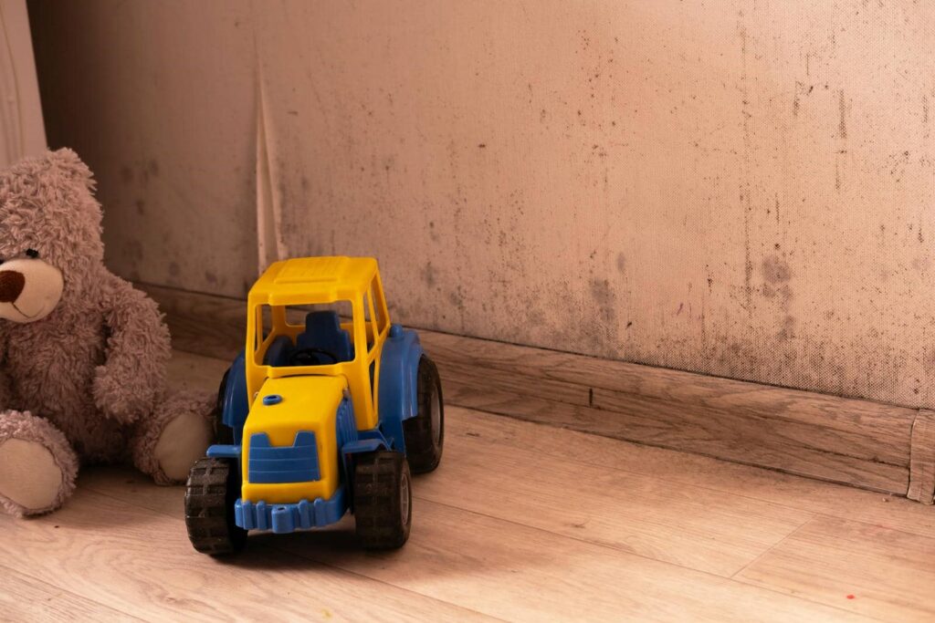 A yellow and blue toy tractor next to a brown teddy bear on a wooden floor with a dirty wall background.