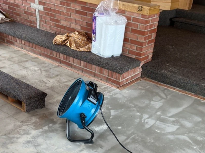 Blue industrial fan on a concrete floor near carpeted brick steps with a plastic-wrapped package on top. trusted restoration company