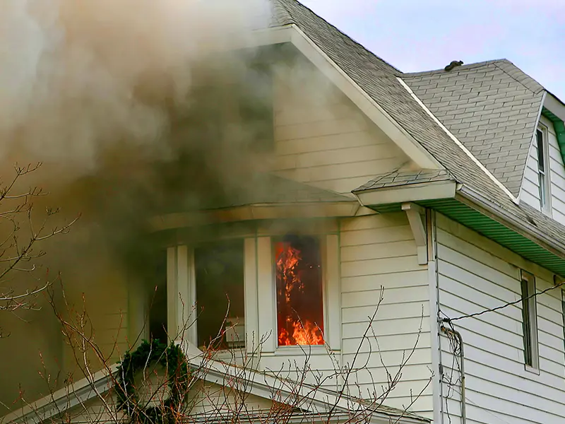 Fire and thick smoke coming from the second-floor window of a white house. fire damage restoration company