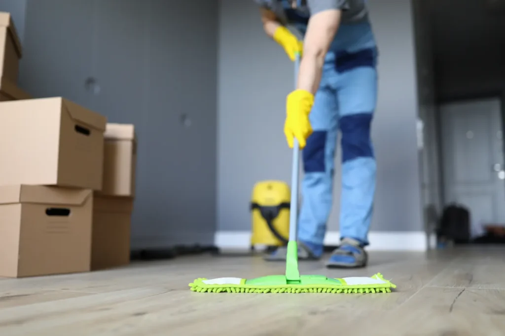 Person wearing yellow gloves mopping a wooden floor near stacked cardboard boxes.