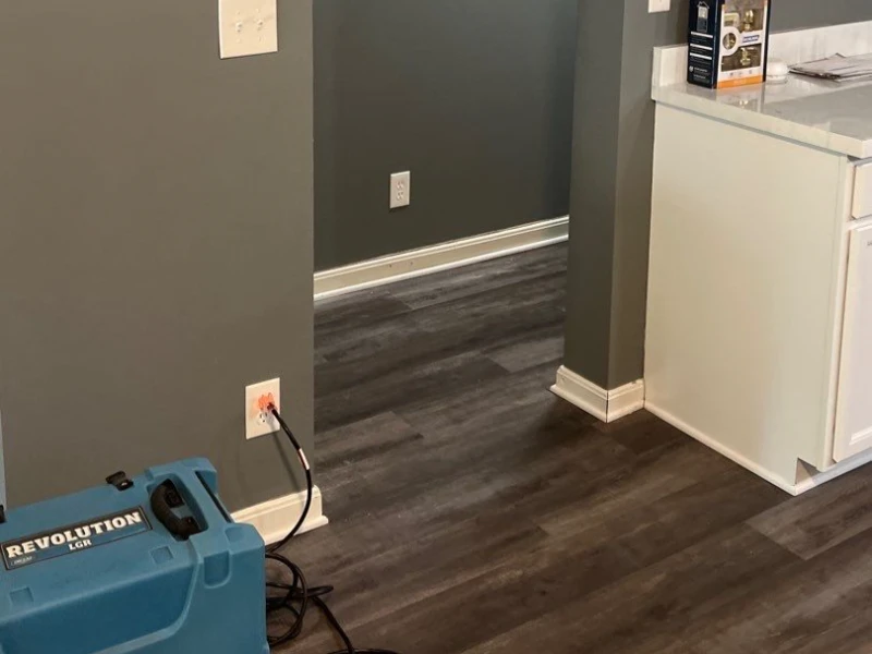 Blue air mover machine plugged into a wall outlet on dark wood flooring near a kitchen counter.